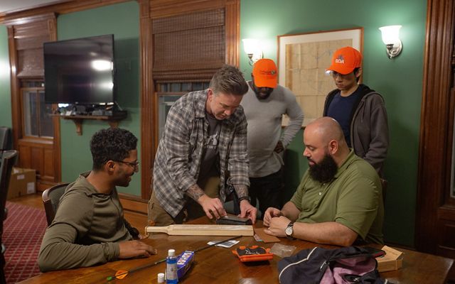 5 men gather around a table in a green room as one holds a small piece of metal in his hand.