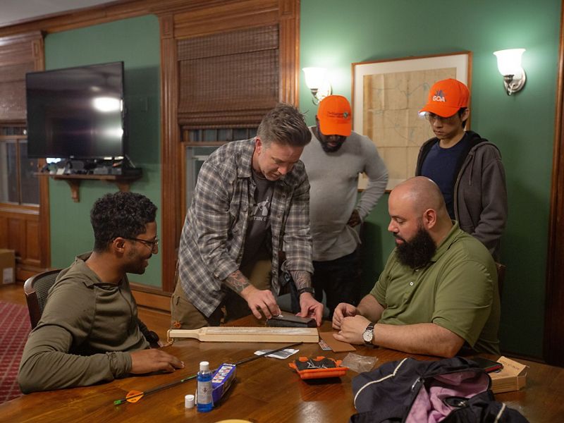 5 men gather around a table in a green room as one holds a small piece of metal in his hand.
