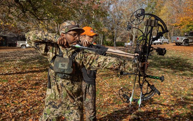 Two men both wearing camo line up arrows on their compound bows.