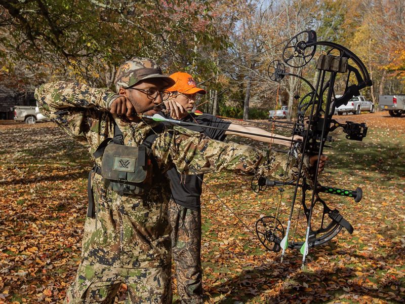 Two men both wearing camo line up arrows on their compound bows.