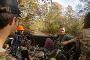 Several hunters ride in the bed of a truck through the woods, holding their gear.