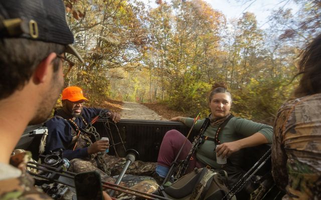 Several hunters ride in the bed of a truck through the woods, holding their gear.