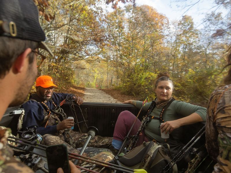 Several hunters ride in the bed of a truck through the woods, holding their gear.