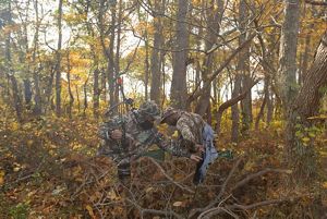 Amid a lot of autumn-colored brush in the woods, one man in camo helps another man in camo put on or take off a jacket.