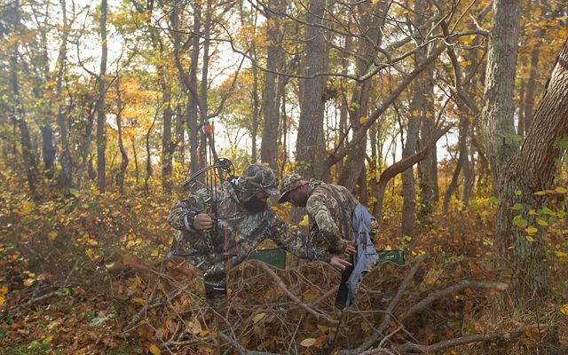 Amid a lot of autumn-colored brush in the woods, one man in camo helps another man in camo put on or take off a jacket.