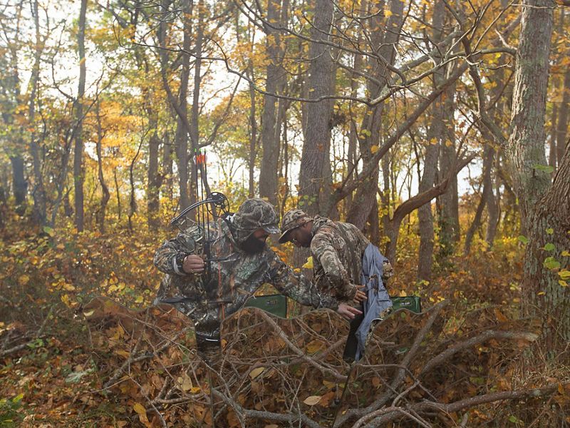 Amid a lot of autumn-colored brush in the woods, one man in camo helps another man in camo put on or take off a jacket.