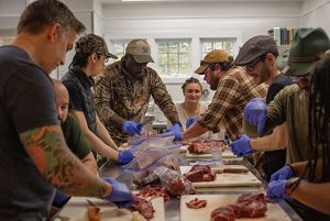 8 adults gather around a long metal table, each with cutting boards, meat and ziploc bags. 