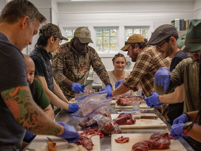 8 adults gather around a long metal table, each with cutting boards, meat and ziploc bags.