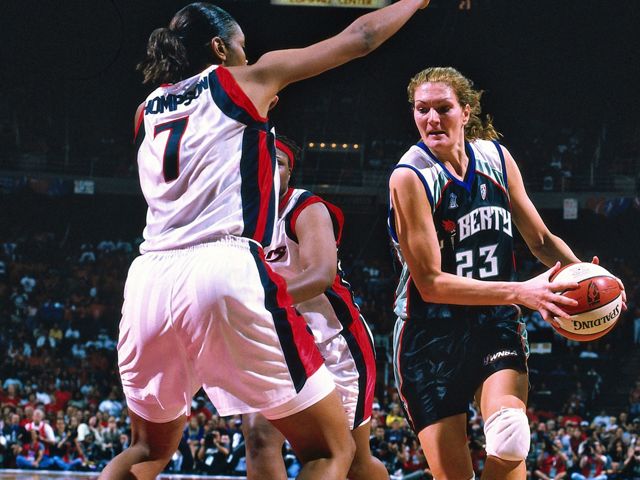 Woman in basketball uniform mid-game with basketball in hand on righthand part of screen.