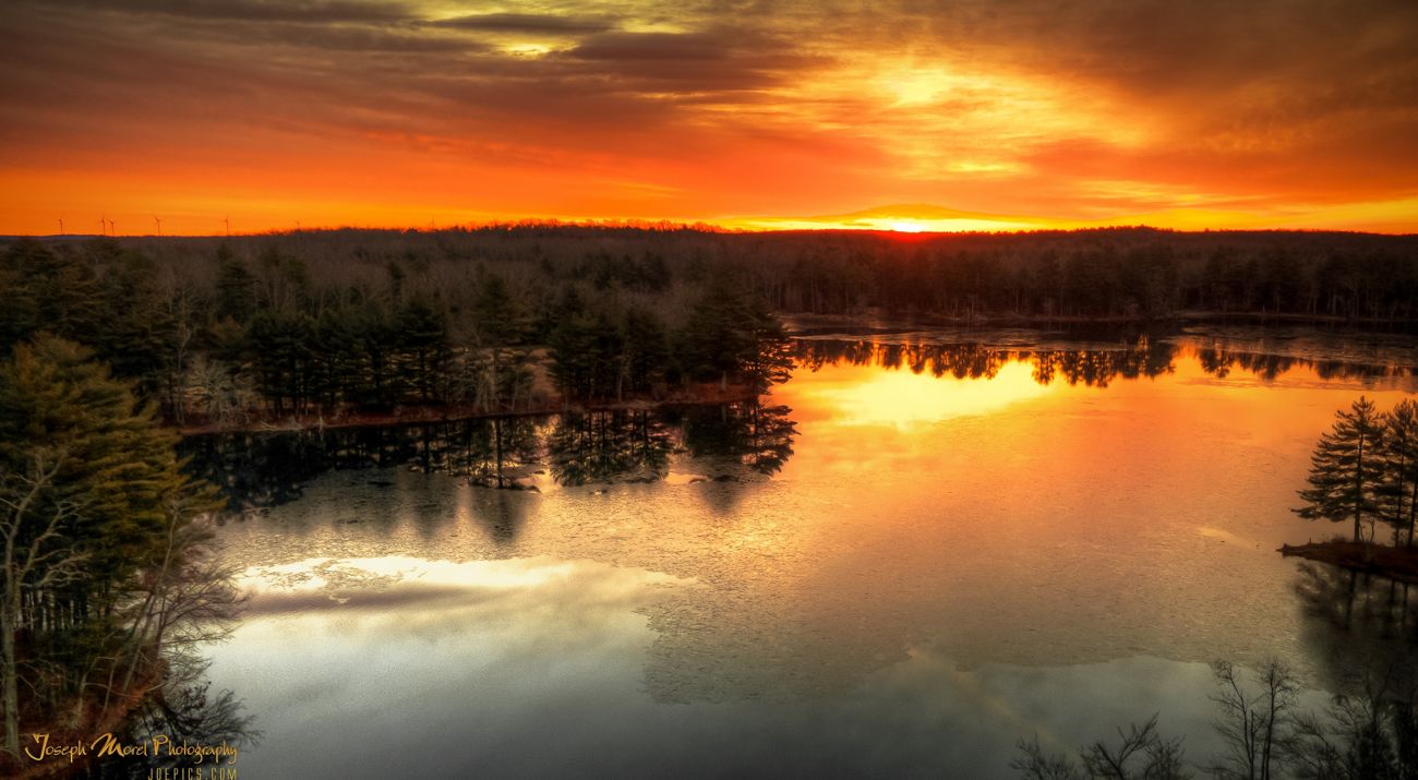 The sun breaks over the horizon and orange skies are reflected in a pond below, ringed by pine trees.