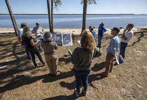 A group of people stand in a semi-circle around a table with maps and information set up on a beach in the shade of two trees.