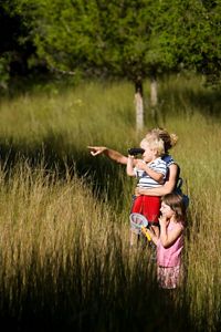 A woman holds up a young boy who is looking through binoculars. A girl holding a bug net stands close by. 