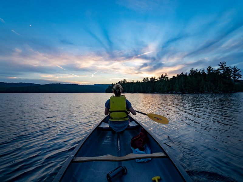 A view from behind a woman paddling a canoe across a lake tinted purple in the sunset.