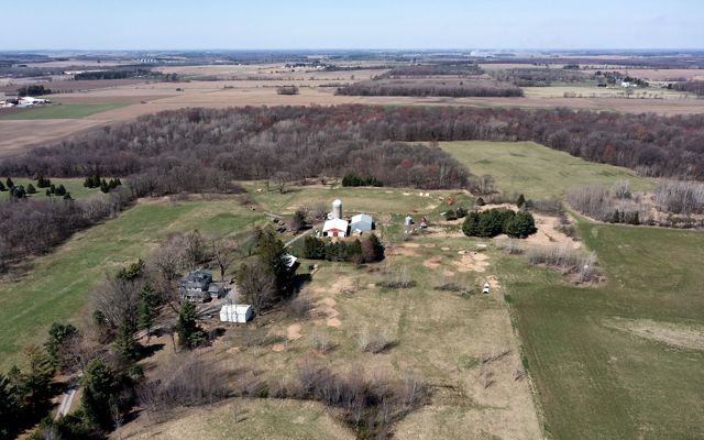 Aerial view of several farm buildings and a silo in the midst of an expansive landscape of green fields and brown-colored woods.