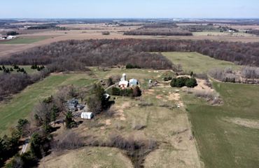 Aerial view of several farm buildings and a silo in the midst of an expansive landscape of green fields and brown-colored woods.