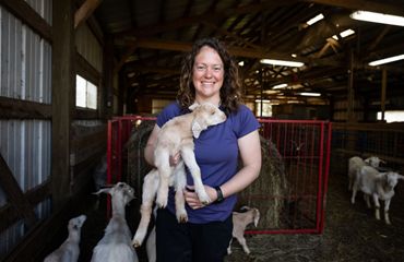Portrait of Leslie Svacina holding a baby goat and standing in a barn with several other goats walking around her.