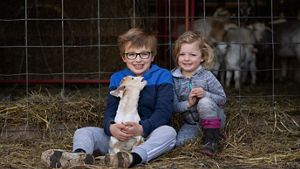 Children Cody and Cady Svacina sit next to each other on hay on the ground in front of a goat barn; Cody holds a baby goat in his lap.