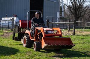 Scott Svacina drives a small orange tractor in front of a farm building.
