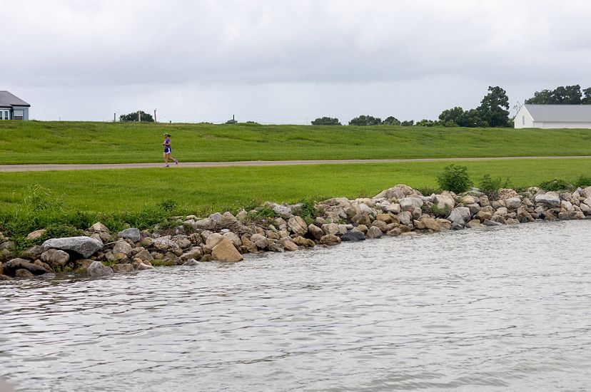 A shot of the Metairie waterfront. Rocks are piled on the shore to protect against waves and storms. A jogger runs down a pathway in front the tall grassy levee. 