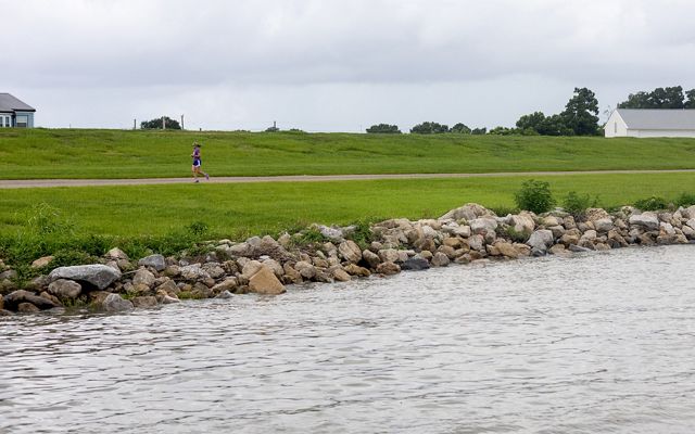 A shot of the Metairie waterfront. Rocks are piled on the shore to protect against waves and storms. A jogger runs down a pathway in front the tall grassy levee. 