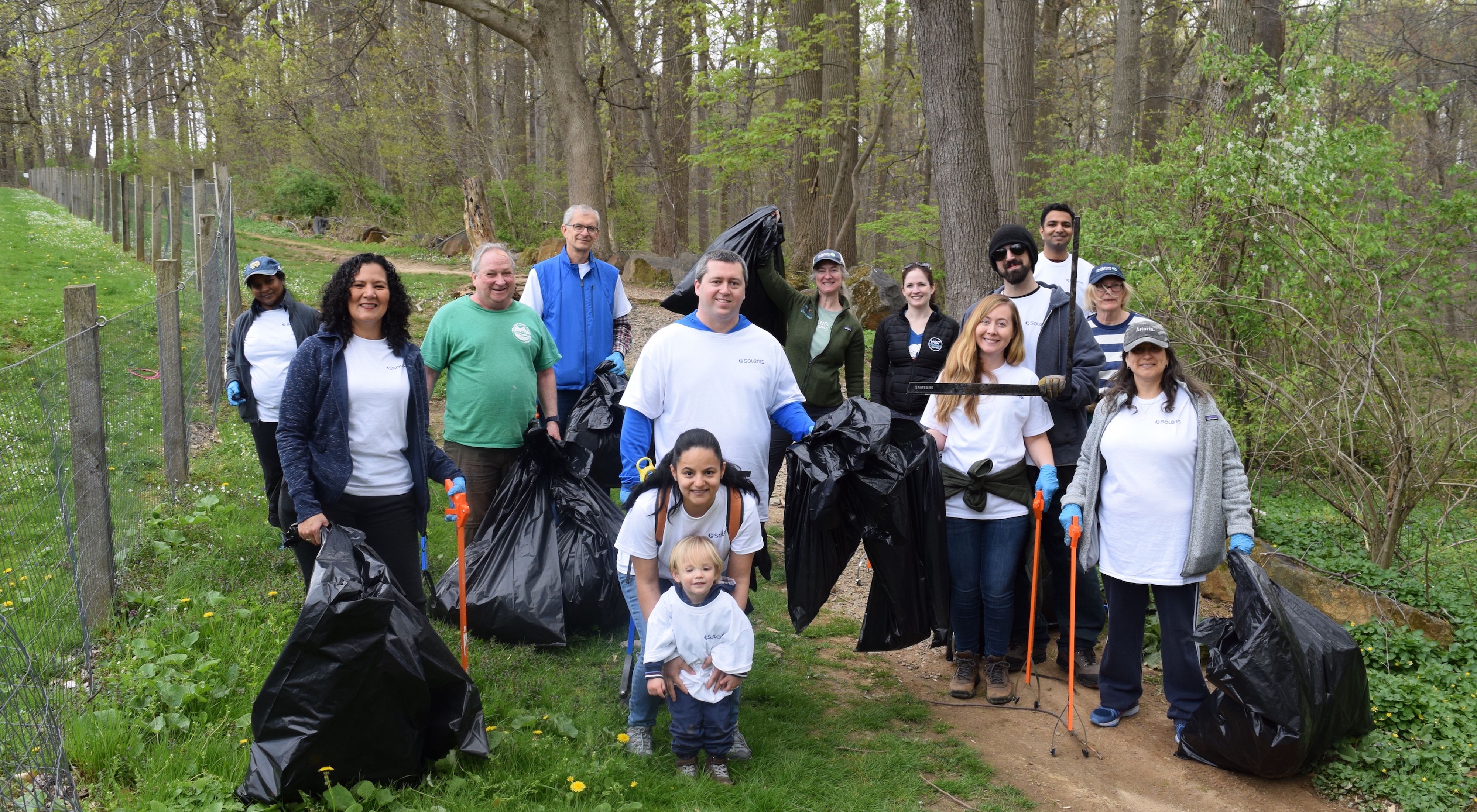 A group of people with trash bags and trash pickers stand on a trail in the forest.