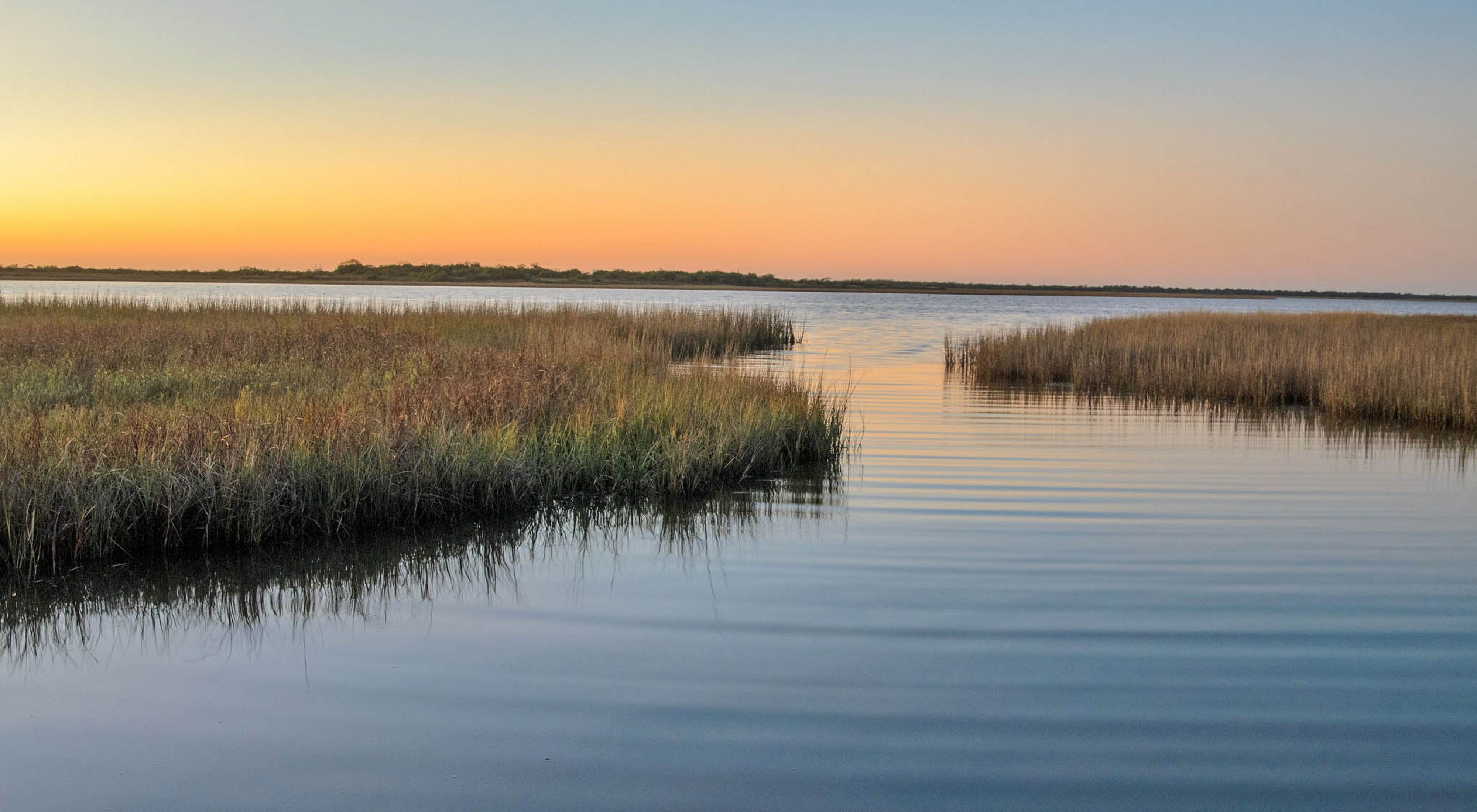 An expanse of dense coastal wetlands intermixed with open water along the Texas Gulf Coast at sunset; the sky is orange.
