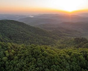 The sun rises over a forested mountain range. Rolling green ridges dominate the foreground and roll away to the horizon. White mist rises from the valleys in the distance.