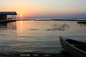 A view of a sunset over water. A dock with a picnic table can be seen on the left with a canoe in the bottom right.