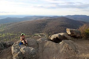 A hiker rests on a large rock, looking out at a beautiful mountain view.