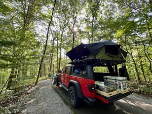 A jeep with a rooftop tent and camping materials in the trunk, parked in a forest.
