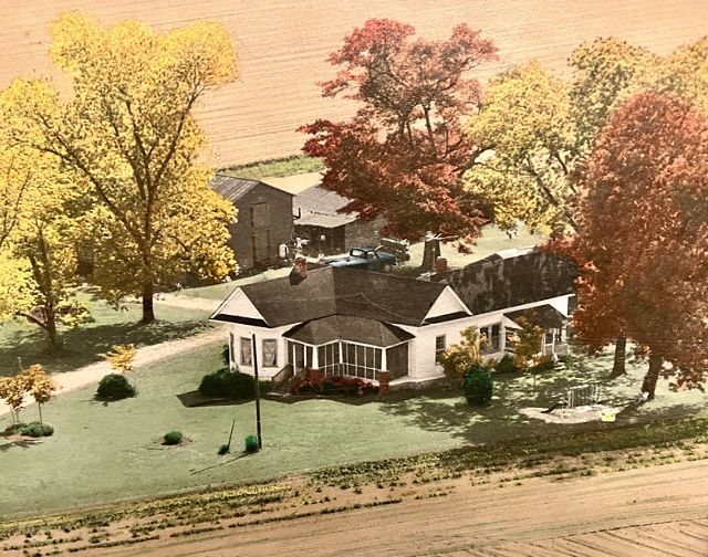 A white farmhouse with a black roof. Behind the house are two barns and the house is surrounded by colorful trees.