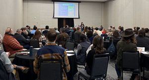A room full of people sit and watch a woman make a presentation.