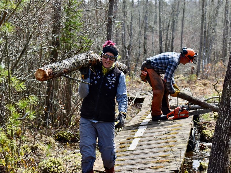 Elizabeth Hanson carries a large log over her shoulder as she walks on a wooden boardwalk.