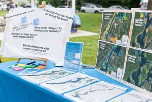 A table with a bright blue tablecloth that has several poster boards on it, one asking visitors questions about flooding, others with flood maps of Taunton, MA.