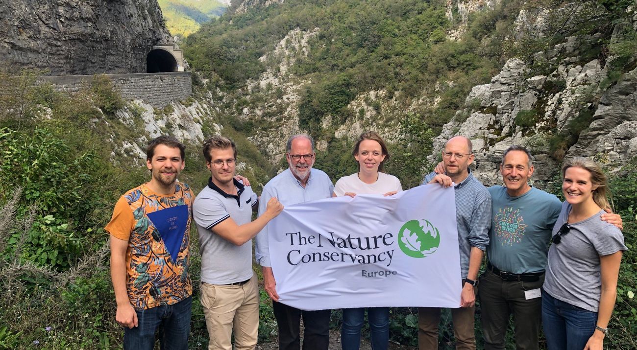Seven people stand together holding a cloth banner with TNC's logo on it with a rocky gorge in the background.