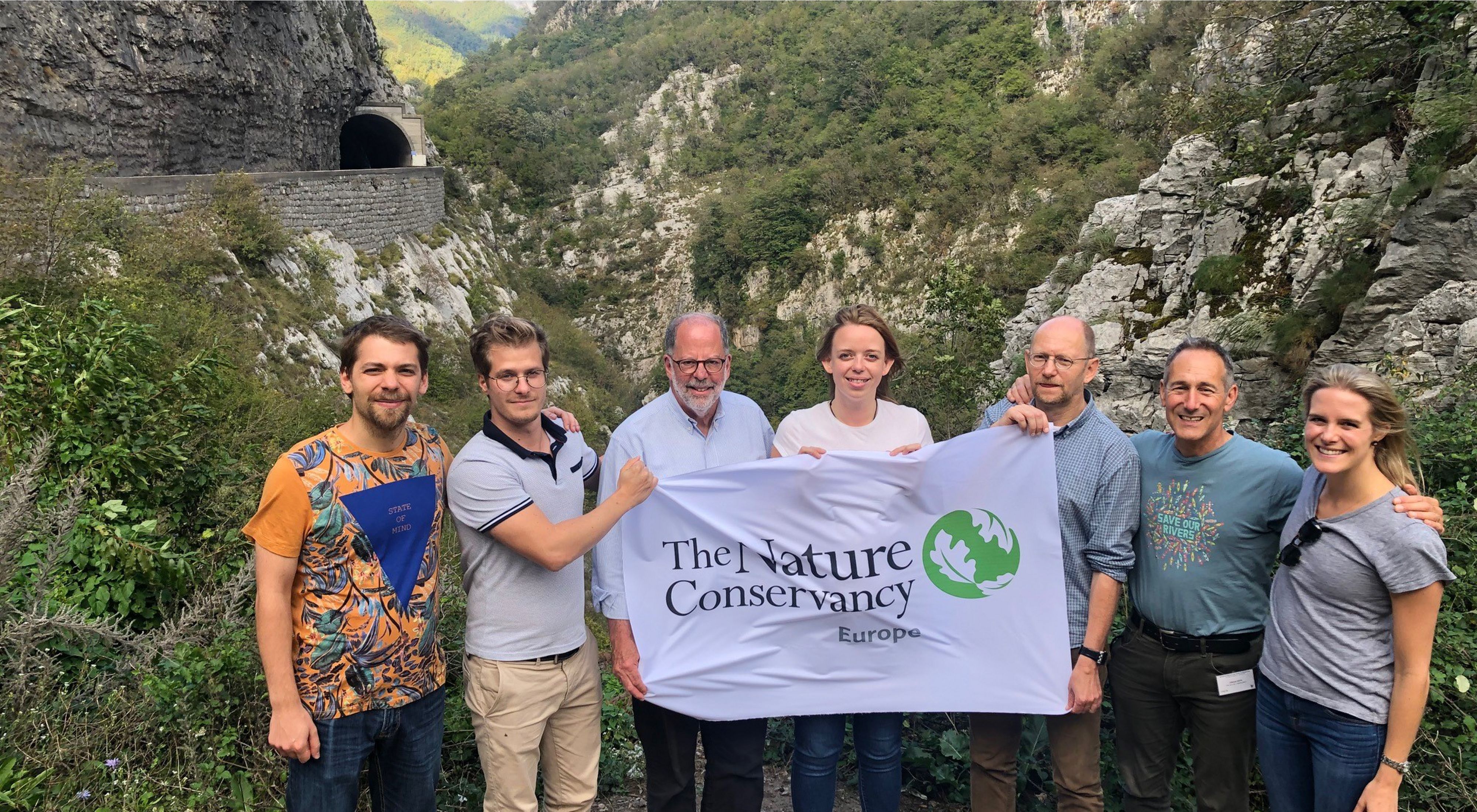 Seven people stand together holding a cloth banner with TNC's logo on it with a rocky gorge in the background.