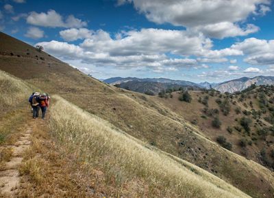 Hikers follow a grassy mountainside path.