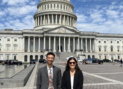 Two smiling people in suits stand in front of the United States Capitol building.