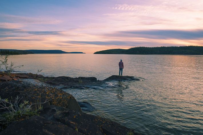 A person stands on a large piece of rock jutting out into the water in Thaidene Nene.