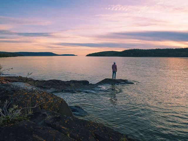 A person stands on a large piece of rock jutting out into the water in Thaidene Nene.