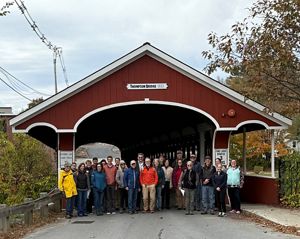 A group of people pose in front of a red wooden covered bridge.