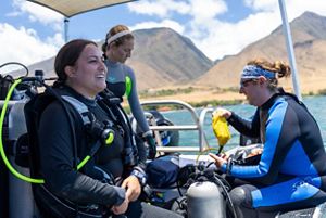 Three people in scuba gear sit on a boat and prepare to dive into the ocean.