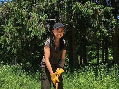 An intern stands among logs while holding a large mallet.