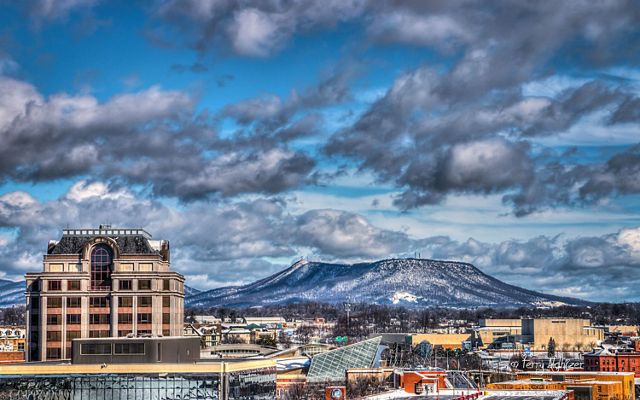 Heavy white clouds hang low over a flat-topped mountain frosted with snow that rises behind the city skyline of Roanoke, VA.