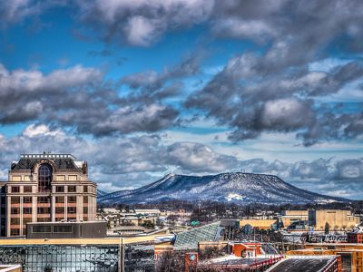 Heavy white clouds hang low over a flat-topped mountain frosted with snow that rises behind the city skyline of Roanoke, VA.