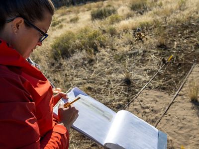 A woman in a red jacket stands above a patch of dirt and writes in a notebook she holds in her hands.