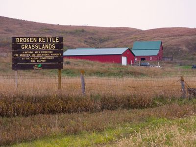 A TNC preserve sign for Broken Kettle Grasslands stands in tall brown grasses with a red barn in the background.