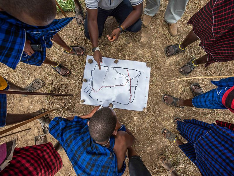 A group of men gather around a small map laid out on the ground.