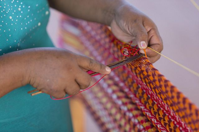a close-up shot frames a person's hand passing a weaving shuttle through a purple, orange and red woven piece