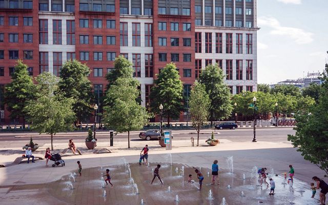 A group of children run and play outdoors, enjoying the water jets at an urban splashpad park in Washington, DC. 